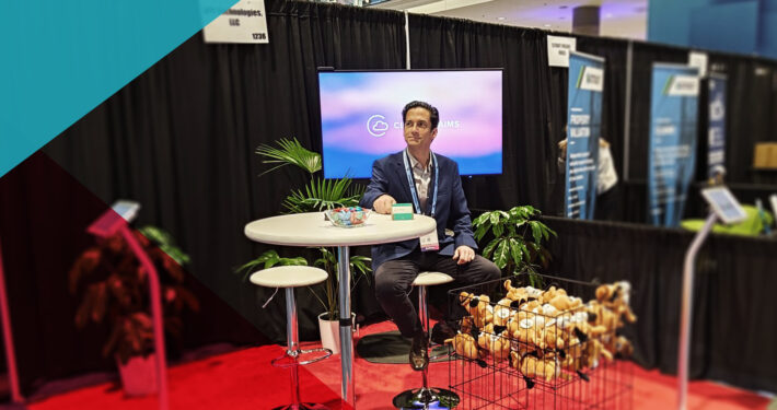 RiskWorld 2025 exhibit hall: Bill Nagle sits at a high-top table branded “Cloud Claims,” beside a wire basket overflowing with “This Is Fine” plush dogs and APPTech materials.