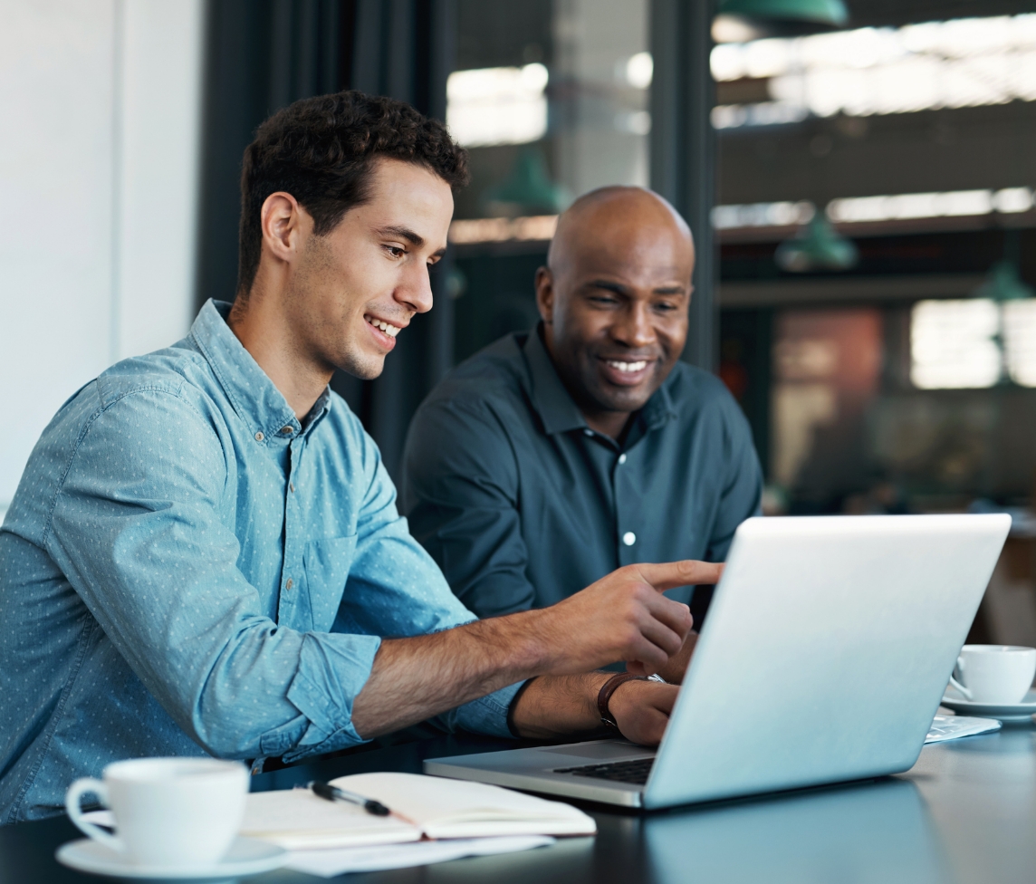 two men sit at a table looking at a laptop